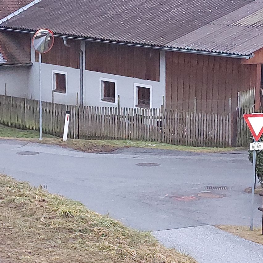 Ein kleines Haus mit braunem Dach und weißen Wänden, umgeben von einem Holzzaun. Ein Kreisverkehr-Schild befindet sich auf der rechten Straßenseite.