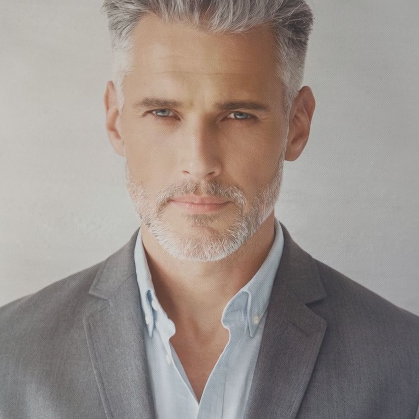A man with gray hair and a beard is wearing a gray suit and a light blue shirt. He is looking straight ahead with a serious expression.
