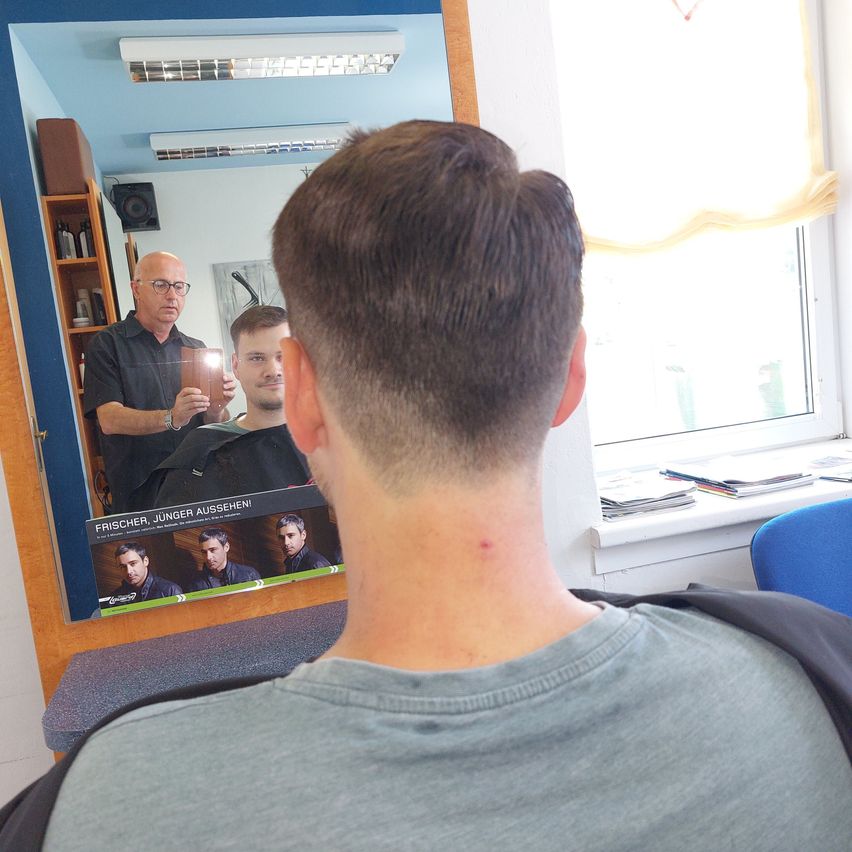 Ein Mann lässt sich in einem Friseursalon die Haare schneiden, reflektiert im Spiegel mit einem Poster von drei Männern.
