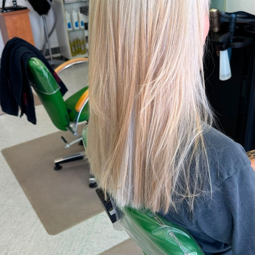 Eine Frau mit langen blonden Haaren sitzt in einem Salonstuhl, mit einem grünen Stuhl und einer schwarzen Tasche hinter ihr.