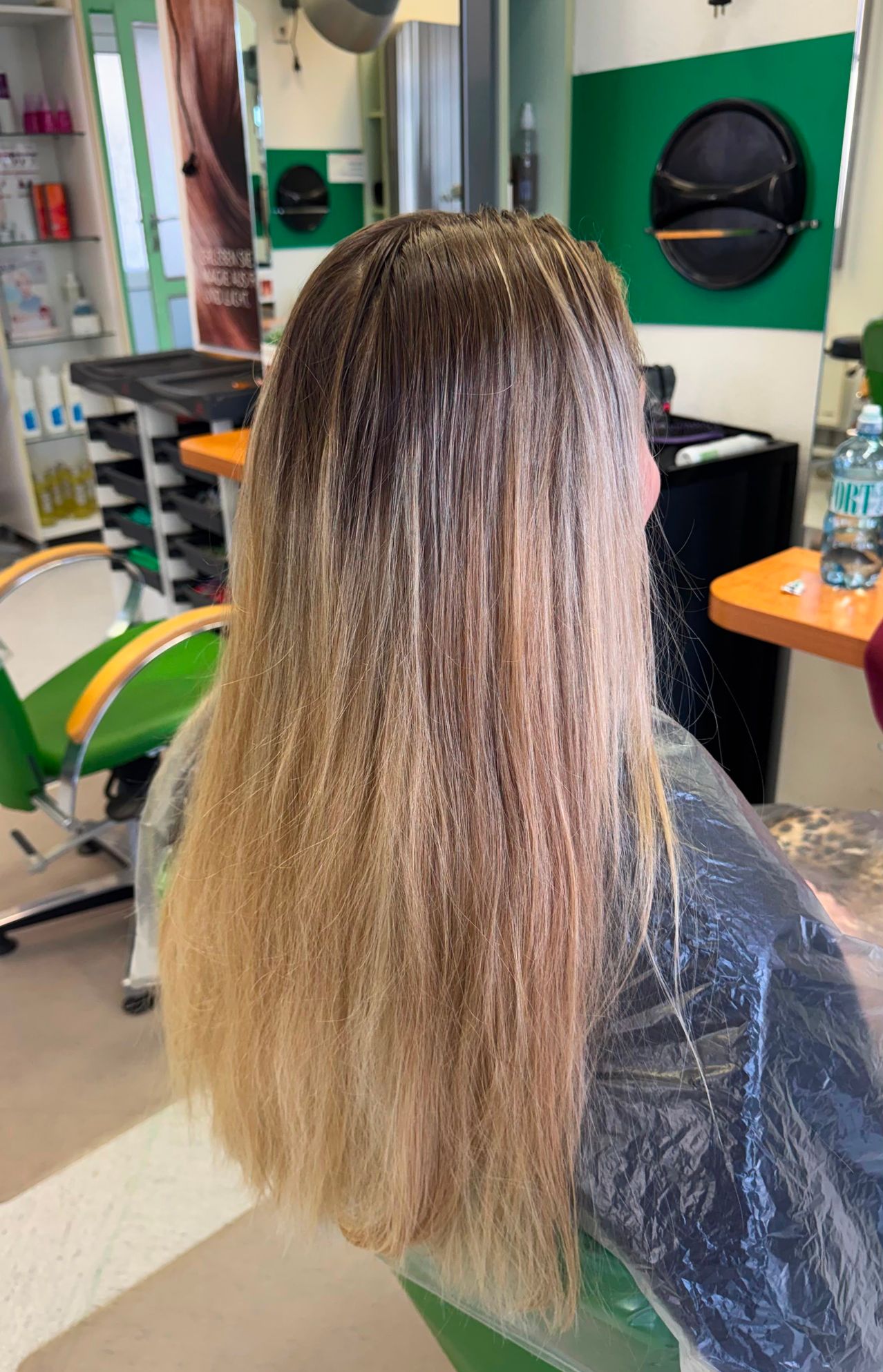 Eine Frau mit langen blonden Haaren sitzt in einem Salonstuhl, ihr Haar ist von einer Plastikschürze bedeckt. Hinter ihr ist ein Schreibtisch mit einer Wasserflasche und einem schwarzen Schrank zu sehen.