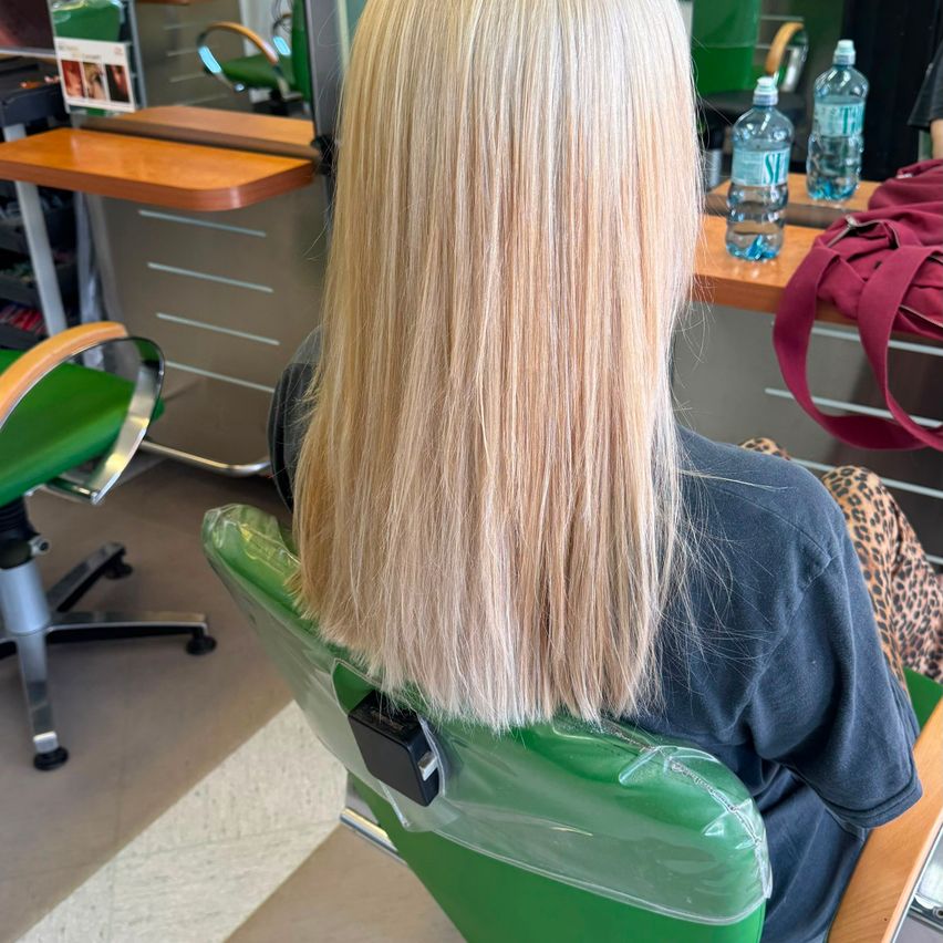 Eine Frau mit langen blonden Haaren sitzt in einem Salonstuhl, mit einem Schreibtisch und Flaschen hinter ihr.