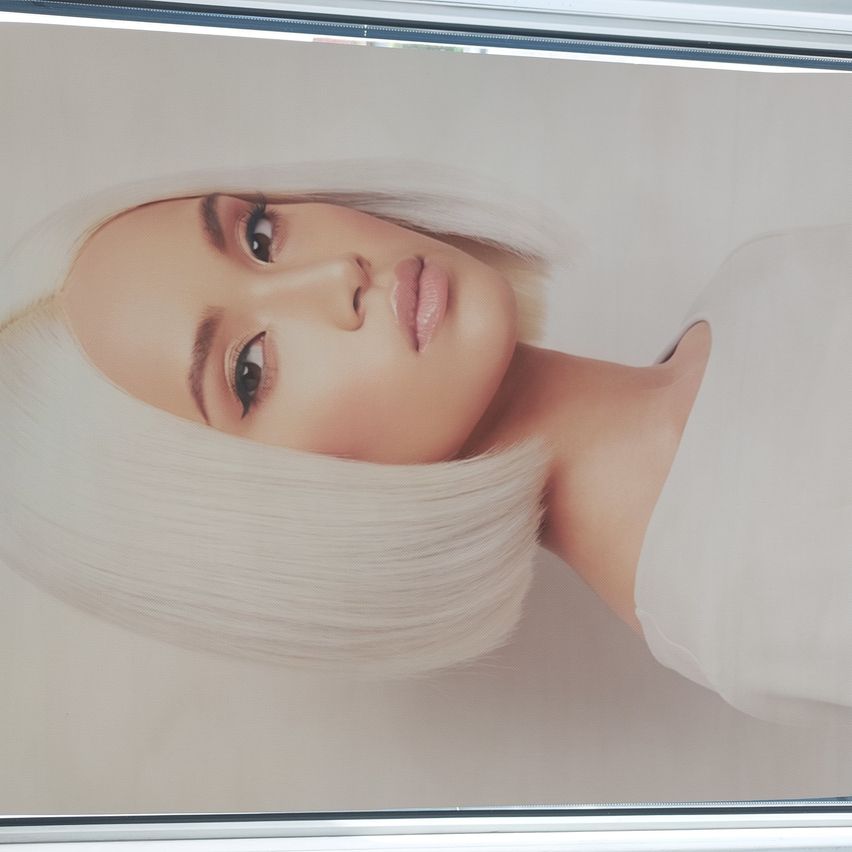 Eine Frau mit platinblondem Haar in einem Bob-Schnitt, das direkt in die Kamera schaut. Sie trägt ein weißes Oberteil, mit leicht geöffneten Lippen und leicht geschlossenen Augen. Das Bild hat einen leichten Unschärfeeffekt.