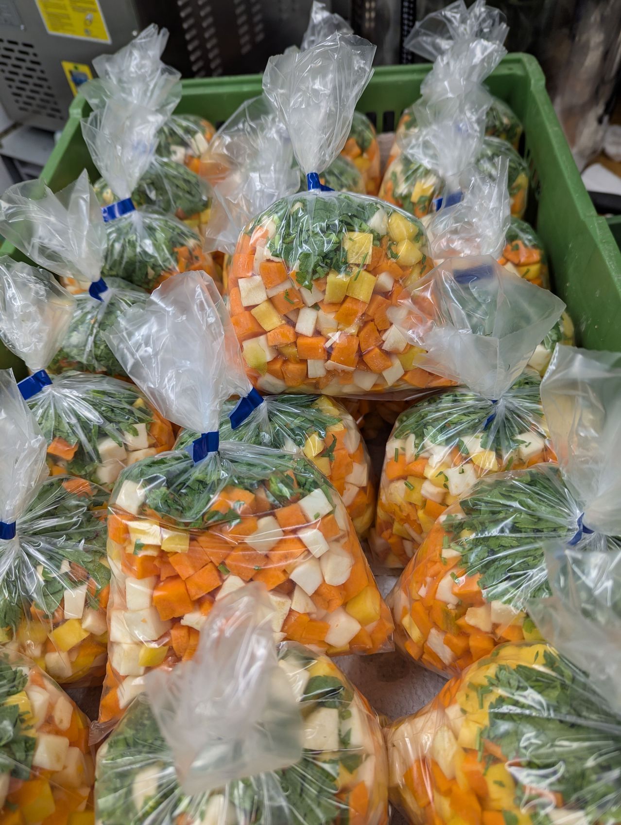 Mehrere durchsichtige Plastiktüten mit gehackten Gemüse, einschließlich Karotten, Sellerie und grünen Kräutern, sind in einem grünen Korb platziert. Jede Tüte ist mit einem blauen Faden gebunden.