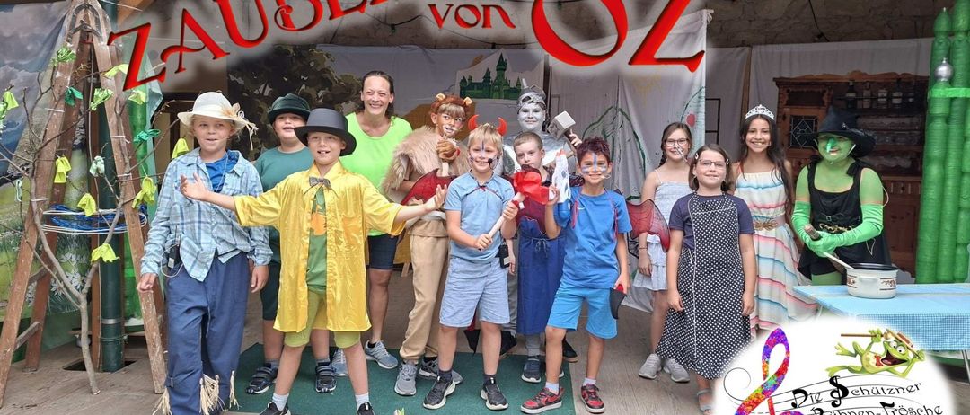 Eine Gruppe von Kindern in verschiedenen Kostümen, darunter Hexen, Teufel und andere, posiert für ein Foto auf einer Bühne mit Blumen und einem Banner mit der Aufschrift 'Der Zauberer von Oz'.
