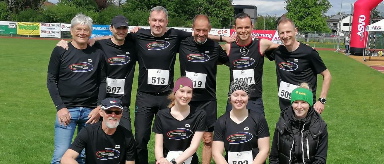 Eine Gruppe von Marathonläufern, sowohl Männer als auch Frauen, posiert für ein Foto auf einem Rasenfeld. Sie tragen alle schwarze T-Shirts und Shorts, wobei ihre Nummern sichtbar sind.
