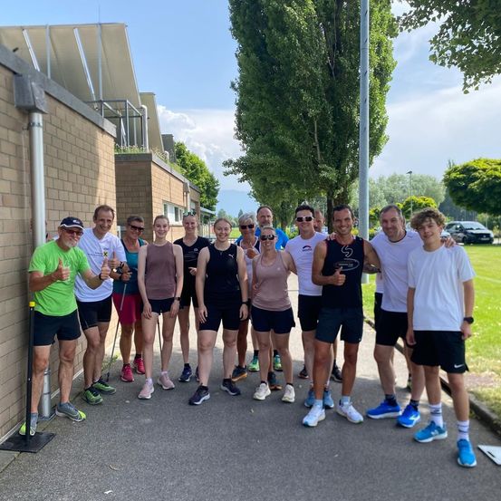 Eine Gruppe von Menschen in Sportbekleidung posiert an einem sonnigen Tag für ein Foto, mit einem Gebäude und Bäumen im Hintergrund.