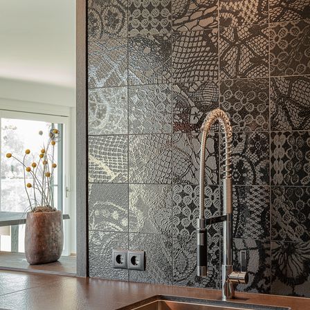 Eine Küche mit einer gefliesten Wand mit Schlangenmuster, einem Waschbecken mit Wasserhahn und einem Topf mit gelben Blumen im Fensterreflex.