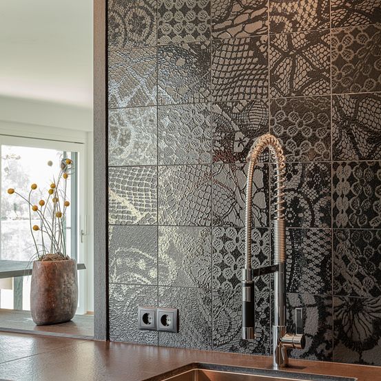 Eine Küche mit einer gefliesten Wand mit Schlangenmuster, einem Waschbecken mit Wasserhahn und einem Topf mit gelben Blumen im Fensterreflex.