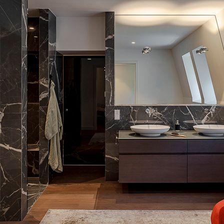 Modernes Badezimmer mit Marmorwänden, zwei Waschbecken und einem großen Spiegel über dem Waschtisch. Ein Handtuch hängt an einem Ständer und ein roter Stuhl steht in der Nähe.