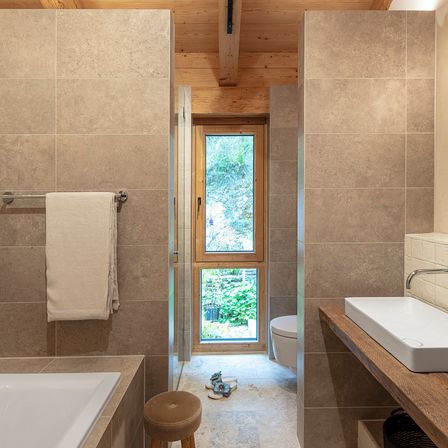 Ein modernes Badezimmer mit beigefarbenen Fliesen, einer Badewanne, einem Fenster, einem WC, einem Waschbecken und einem Handtuch an der Wand.