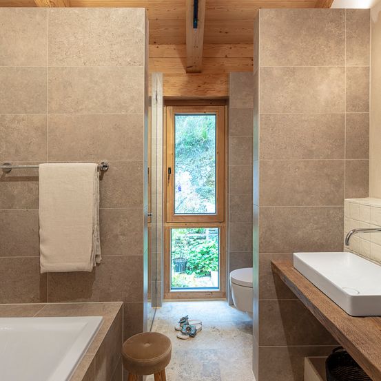 Ein modernes Badezimmer mit beigefarbenen Fliesen, einer Badewanne, einem Fenster, einem WC, einem Waschbecken und einem Handtuch an der Wand.