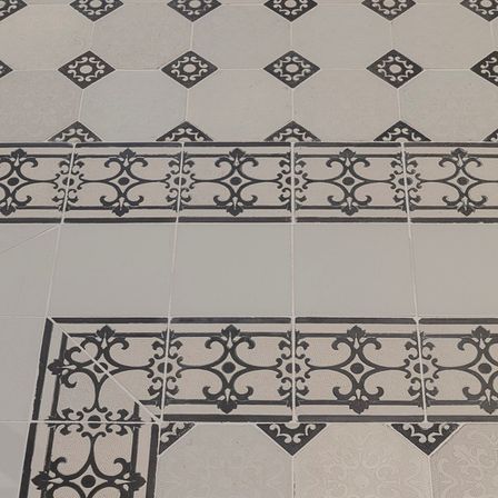 Eine Nahaufnahme eines gefliesten Bodens mit einem sechseckigen Muster. Die Fliesen haben komplizierte schwarze Designs und sind in einem Raster angeordnet. Die Fliesen sind hell und die schwarzen Designs sorgen für Kontrast.