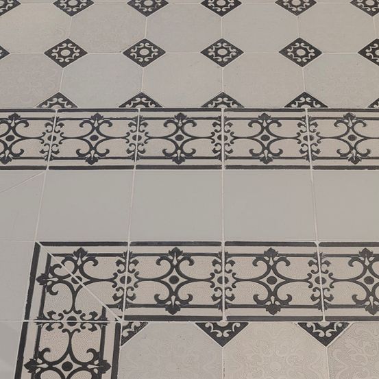 Eine Nahaufnahme eines gefliesten Bodens mit einem sechseckigen Muster. Die Fliesen haben komplizierte schwarze Designs und sind in einem Raster angeordnet. Die Fliesen sind hell und die schwarzen Designs sorgen für Kontrast.