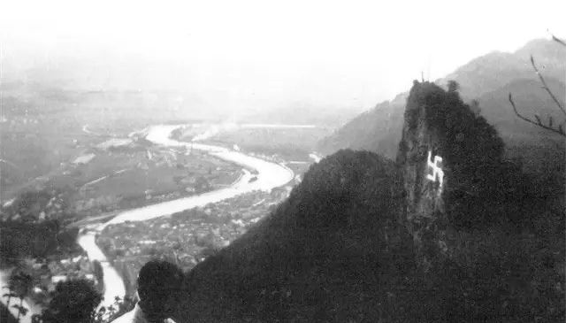 Schwarz-Weiß-Foto eines Mannes, der einen Tal mit einem Fluss und einem Berg betrachtet. Ein Nazi-Symbol befindet sich auf dem Berg.