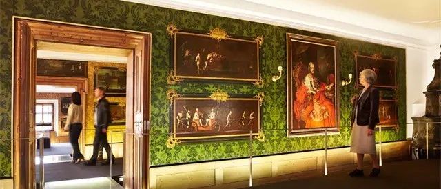 Eine Frau betrachtet eine Wand mit Gemälden in einem Museum, während zwei Personen im Türrahmen hinter ihr stehen. Der Raum hat eine grün tapezierte Wand und einen schwarzen Teppichboden.