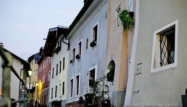 Eine schmale Straße mit mehrfarbigen Gebäuden, einige mit Fenstern, Balkonen und Topfpflanzen. Eine Person geht auf dem Bürgersteig, und Straßenlaternen säumen die Straße.