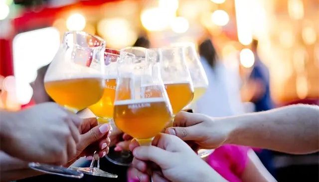 Eine Gruppe von Menschen, die Weingläser mit gelber Flüssigkeit, möglicherweise Bier, in einem Innenraum halten, wahrscheinlich in einem Restaurant, mit verschwommenen Lichtern und Menschen im Hintergrund.