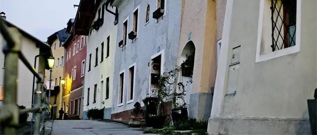Eine schmale Straße gesäumt von mehreren Häusern, jedes mit unterschiedlichen Farben und architektonischen Merkmalen. An den Fenstern hängen Topfpflanzen und eine Laterne beleuchtet den Weg.