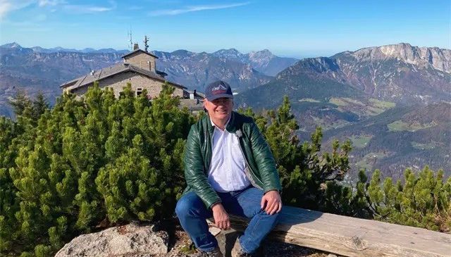 Ein Mann sitzt auf einer Holzbank auf einem Berg mit Blick auf eine Bergkette und ein Gebäude in der Ferne.