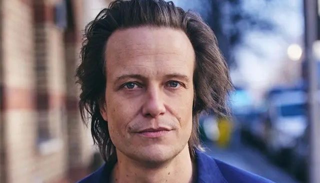 Ein Mann mit langen, glatten Haaren, trägt einen blauen Anzug und steht vor einem Gebäude mit geparkten Autos.