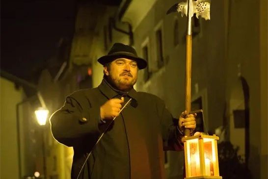 Ein Mann in mittelalterlicher Kleidung hält eine Lanze und eine Laterne in einer schwach beleuchteten Straße bei Nacht.