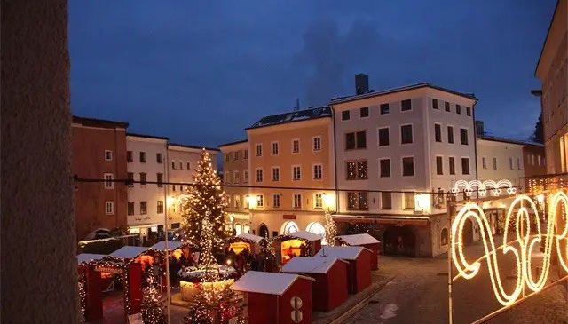 Ein Weihnachtsmarkt ist auf dem Stadtplatz aufgebaut, umgeben von einer Ringreihe roter Hütten. Ein großer Weihnachtsbaum steht in der Mitte, und Gebäude sind im Hintergrund.