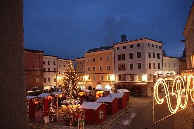 Ein Weihnachtsmarkt ist auf dem Stadtplatz aufgebaut, umgeben von einer Ringreihe roter Hütten. Ein großer Weihnachtsbaum steht in der Mitte, und Gebäude sind im Hintergrund.