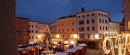 Eine Stadtstraße bei Nacht mit einem Weihnachtsmarkt. In der Mitte steht ein Weihnachtsbaum mit festlichen Lichtern und roten Hütten drumherum. Ein Schild mit hellen gelben Buchstaben buchstabiert das Wort "WEIHNACHTEN".