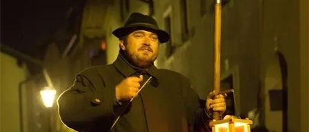 Ein Mann in einem schwarzen Mantel und Hut hält bei Nacht eine Laterne und eine Lanze auf einer Straße.