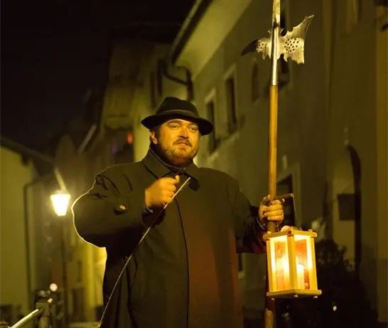 Ein Mann in einem schwarzen Mantel und Hut hält bei Nacht eine Laterne und eine Lanze auf einer Straße.