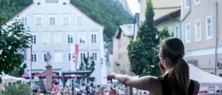 Eine Frau steht auf einer Bühne und deutet auf eine Menge vor einem weißen Gebäude mit vielen Fenstern, vor einem bergigen Hintergrund.