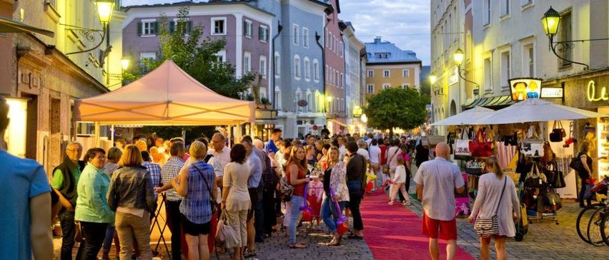 Eine geschäftige Marktstraße in der Dämmerung, mit einer Menschenmenge, die auf einem roten Teppich entlangspaziert, Geschäften mit Markisen und historischen Gebäuden, die die Straße säumen.