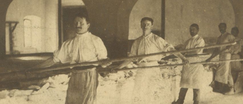 Ein Schwarz-Weiß-Bild zeigt mehrere Männer in Schürzen, die lange Stangen in einem Gebäude tragen. Der Raum hat gewölbte Türen und einen Holzboden.
