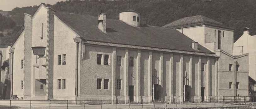 Ein altes Gebäude mit einem Schieferdach und Schornsteinen steht vor einer Steinmauer. Es hat viele Fenster und einen kleinen Turm auf der rechten Seite. Im Hintergrund befindet sich ein Berg mit Bäumen.