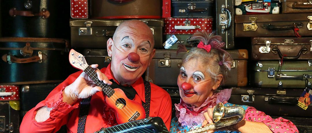 Zwei Clowns, einer hält eine Ukulele und der andere eine Trompete, stehen vor einer Wand voller Koffer.