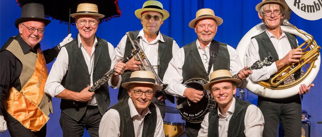 Eine Gruppe von Männern in Vintage-Kleidung, einige mit Hüten, posieren mit verschiedenen Musikinstrumenten, darunter eine Posaune und Trommeln, vor einem blauen Hintergrund.