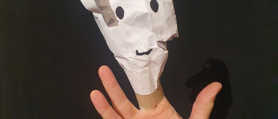 Eine Hand hält eine Papierpuppe mit weißem Gesicht und Ohren vor einem schwarzen Hintergrund.
