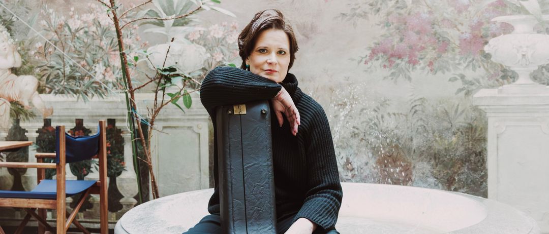 Eine Frau sitzt auf einer runden Steinbank und hält eine schwarze Tasche, mit einem floralen Wandbild im Hintergrund.