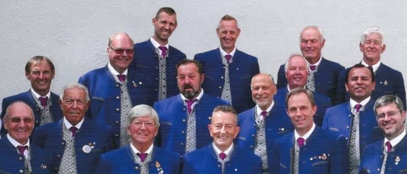 Ein Plakat zeigt eine Gruppe von Männern in blauen Uniformen mit einem roten Hintergrund. Die Gruppe steht vor einer Wand mit dem Wort Kuchl oben. Der Veranstaltungstermin ist der 14. Dezember 2025 um 19:00 Uhr.