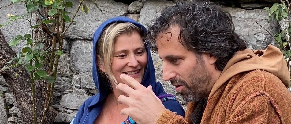 Eine Frau in einem blauen Gewand lächelt, während ein Mann in einem braunen Gewand in der Nähe einer Steinmauer gestikuliert.