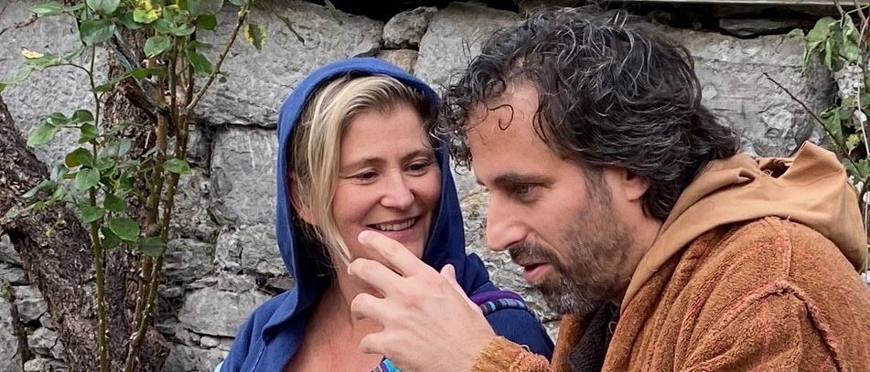 Ein Mann und eine Frau sitzen draußen, die Frau lächelt den Mann an, der zeigt und spricht. Sie tragen Roben, die Frau in Blau und der Mann in Braun. Hinter ihnen steht eine Steinmauer und ein kleiner Baum.