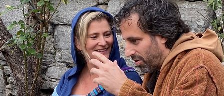 Ein Mann und eine Frau in mittelalterlicher Kleidung sitzen auf einer Steinmauer, lächeln und schauen sich an.