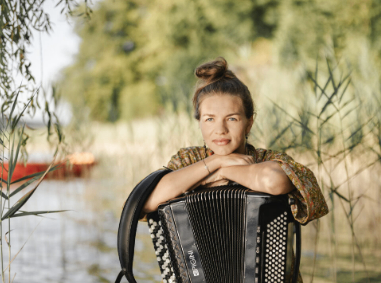 Eine Frau steht am See, lächelt und posiert für ein Foto. Sie hält ein Akkordeon in den Händen und hat einen Rucksack auf dem Rücken. Hinter ihr stehen hohe Gräser und Bäume, und ein Boot ist in der Ferne sichtbar.