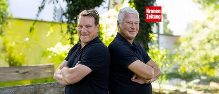 Zwei Männer in schwarzen T-Shirts, einer mit grauen Haaren und einer mit braunen, lächeln und stehen nebeneinander mit verschränkten Armen. Sie stehen vor einer gelben Wand und einem roten Kronen Zeitung-Schild.
