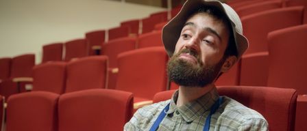 Ein bärtiger Mann in einem blauen Schürze und einer Mütze sitzt in einem roten Stuhl in einem Auditorium.