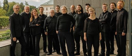 Eine Gruppe von Musikern in schwarzer Kleidung posiert für ein Foto auf einer Terrasse, mit einem grauen Gebäude und bewölktem Himmel im Hintergrund.