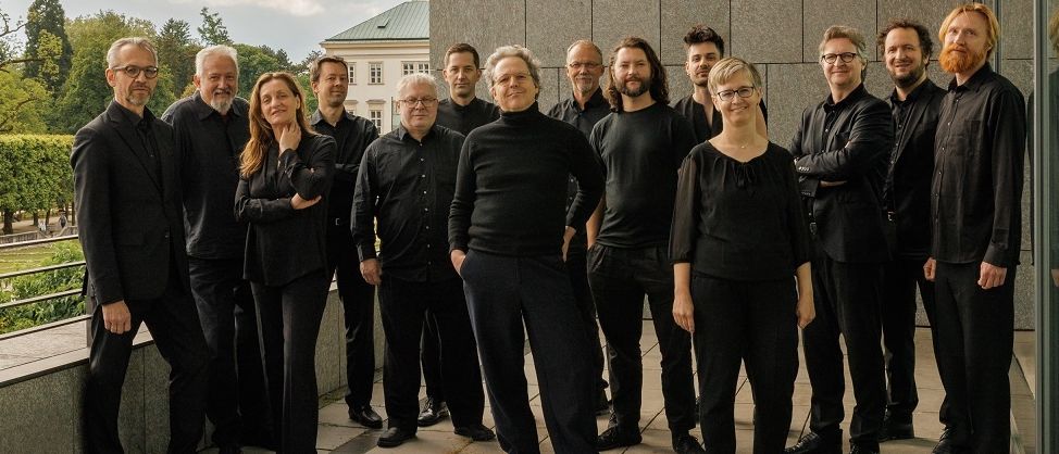 Eine Gruppe von Musikern in schwarzer Kleidung posiert für ein Foto auf einer Terrasse, mit einem grauen Gebäude und bewölktem Himmel im Hintergrund.