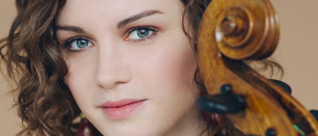 Eine junge Frau mit lockigem Haar lächelt, während sie ein Cello hält. Sie trägt ein schwarzes Oberteil und Ohrringe. Das Cello hat einen braunen Holzkorpus und einen schwarzen Hals.