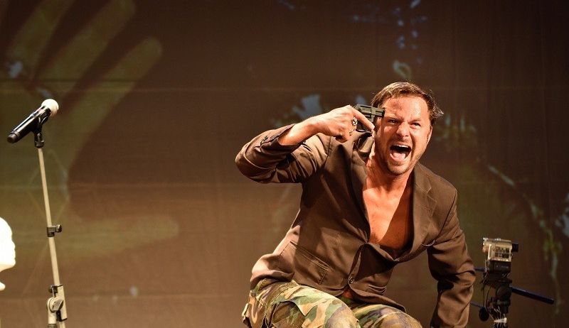 Ein Mann in einem braunen Anzug und Camouflage-Hosen hält eine Waffe auf der Bühne und schreit.